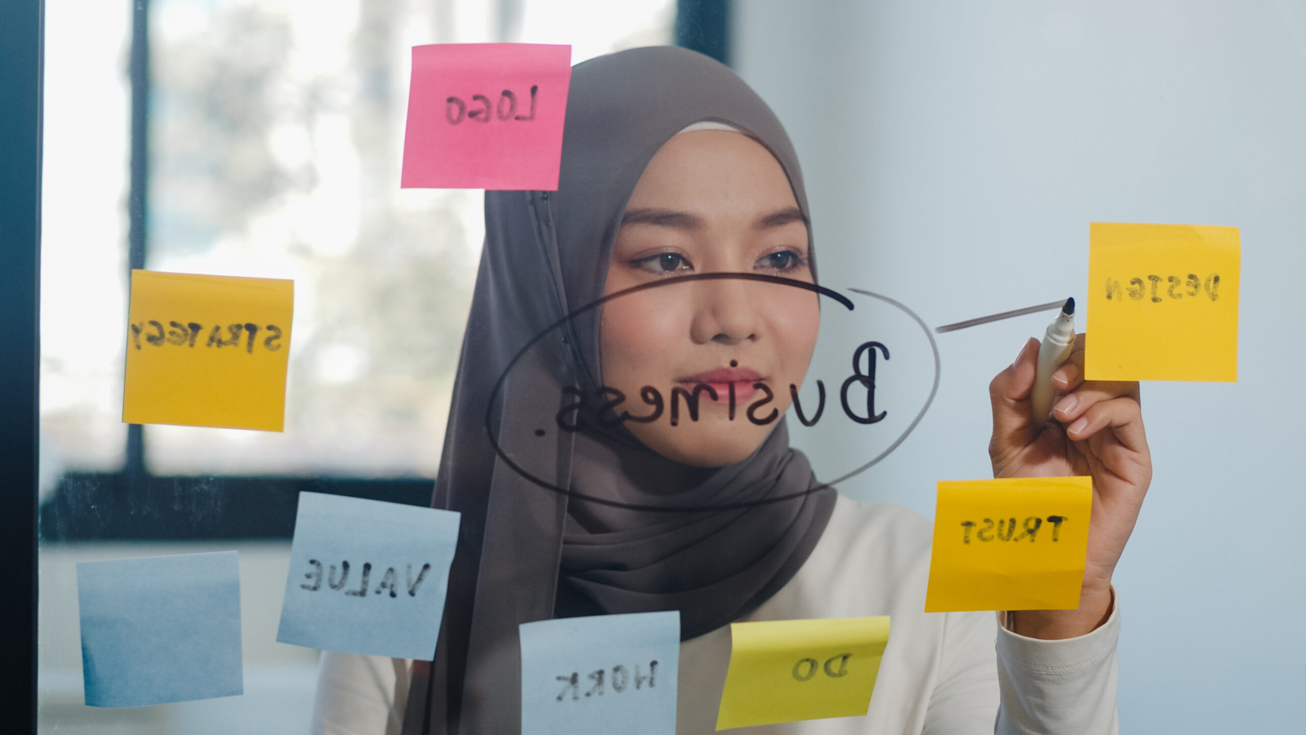 Asia muslim lady write information, strategy, reminder on glass board in new normal office. Working from home, remotely work, self isolation, social distancing, quarantine for coronavirus prevention.