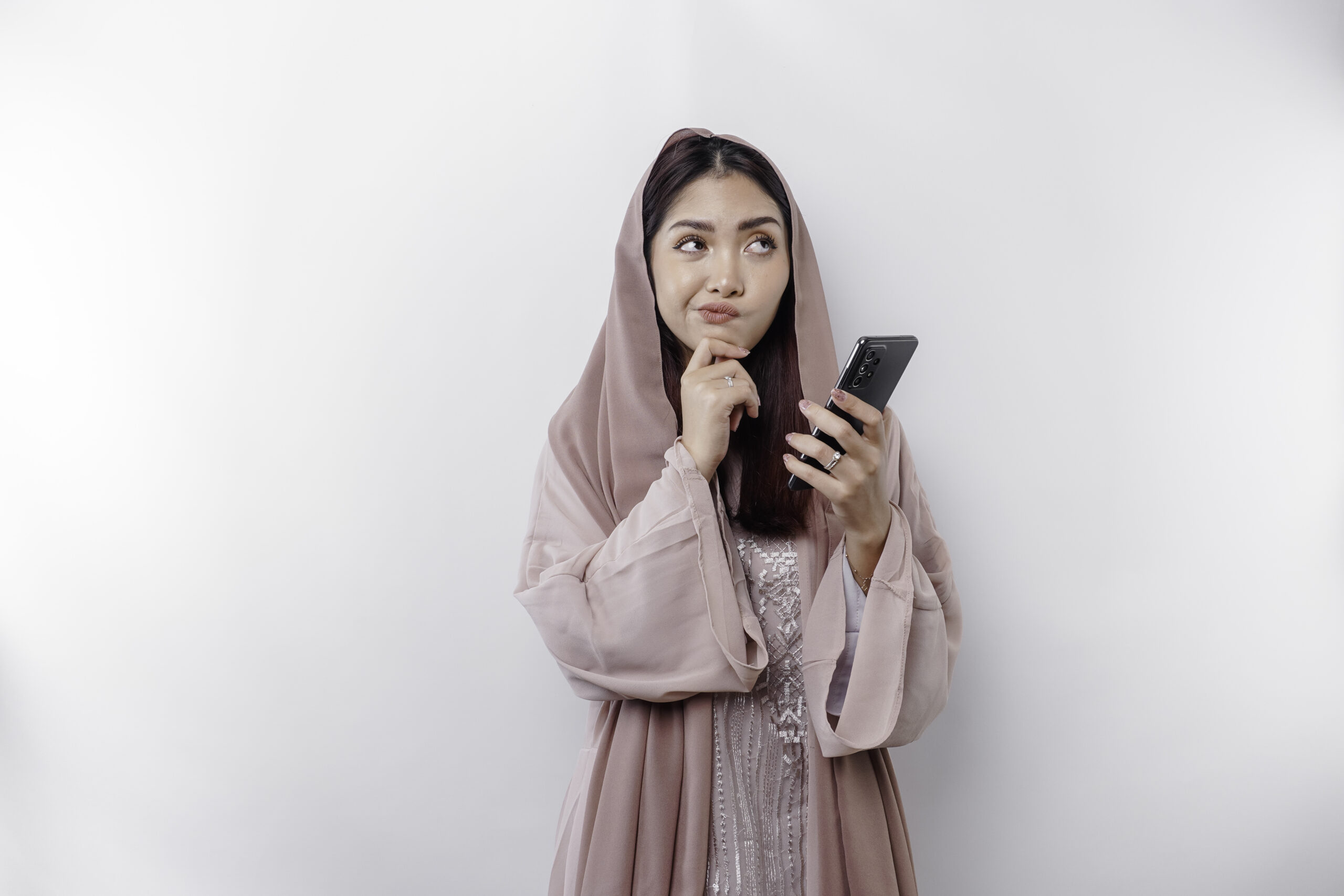 A thoughtful young Asian Muslim woman wearing a hijab and holding her chin while talking on the phone isolated by a white background